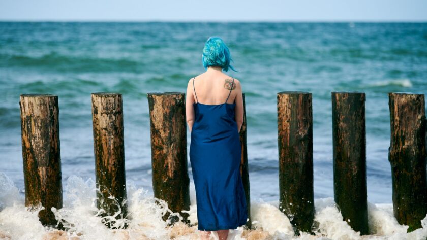 Young blue-haired woman in long dark blue dress standing on sandy beach looking at ocean horizon
